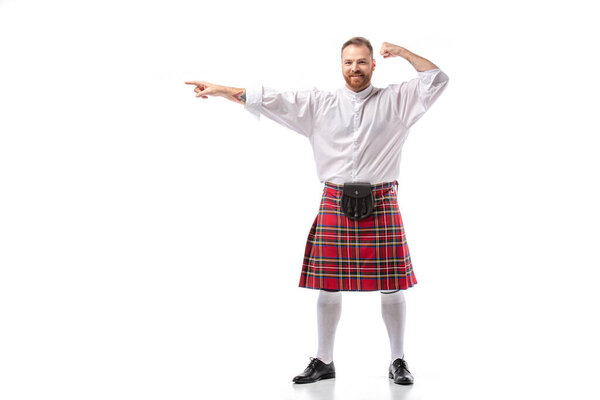 happy Scottish redhead man in red kilt pointing with finger aside on white background