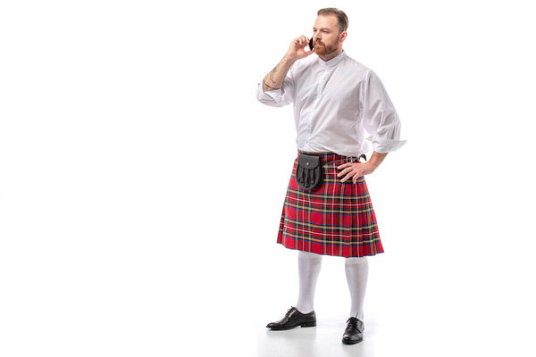 serious Scottish redhead man in red kilt talking on smartphone on white background