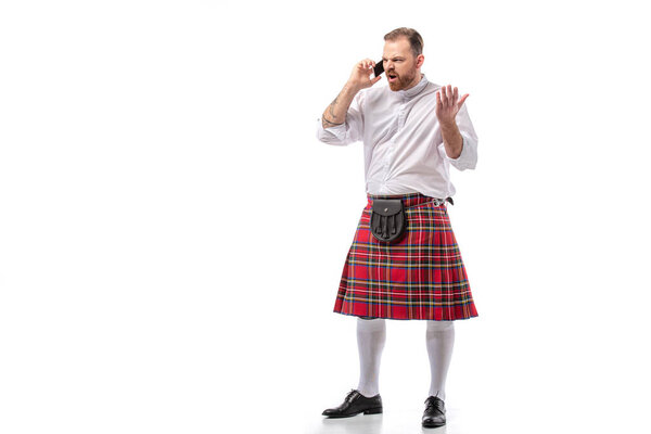 irritated Scottish redhead man in red kilt talking on smartphone on white background