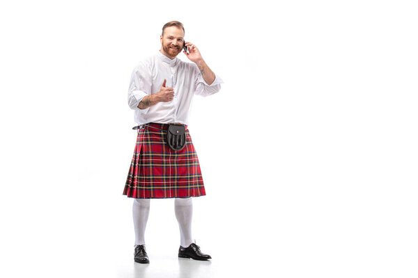 happy Scottish redhead man in red kilt talking on smartphone and showing thumb up on white background