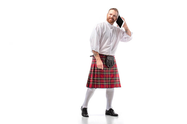 smiling Scottish redhead man in red kilt talking on digital tablet on white background