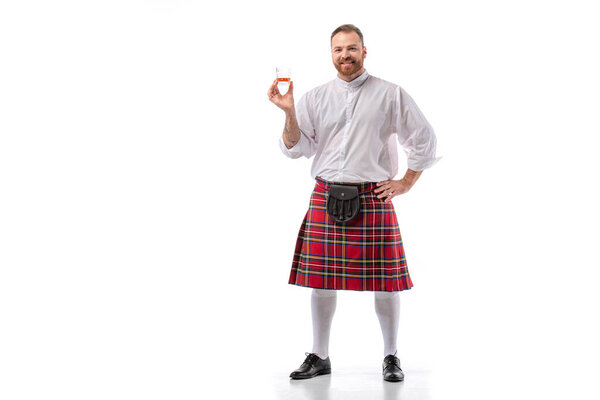 smiling Scottish redhead man in red kilt with whiskey in glass on white background