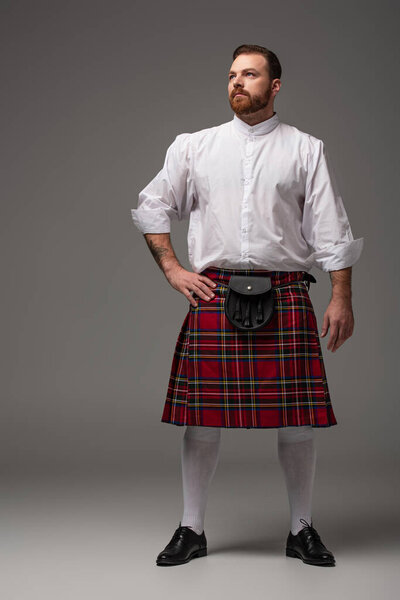 Scottish redhead man in red kilt with hand on hip looking away on grey background