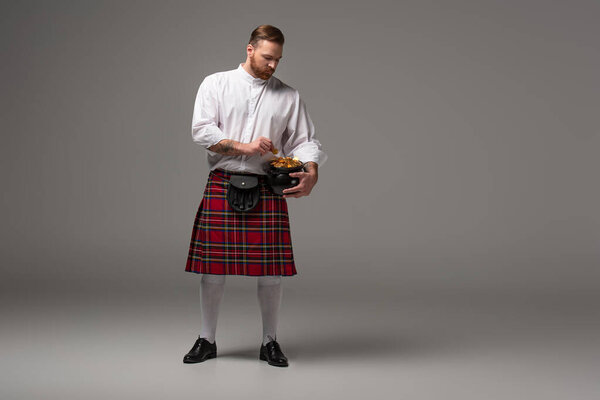 Scottish redhead man in red kilt holding potty with gold coins on grey background