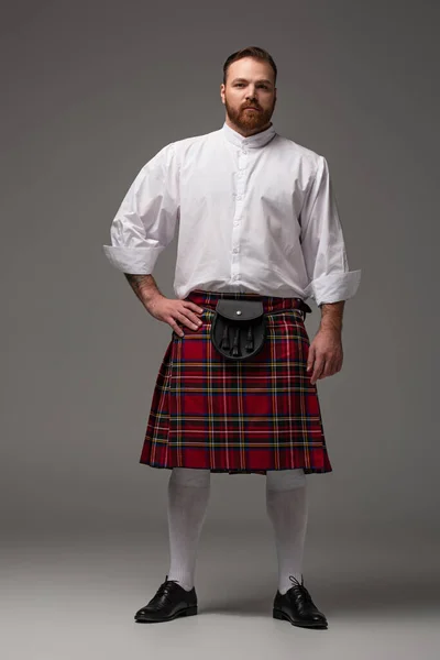 Scottish redhead man in red kilt with hand on hip on grey background