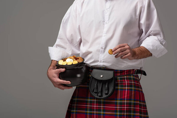 cropped view of Scottish man in red kilt holding potty with gold coins on grey background