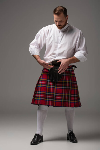 Scottish redhead man in red kilt putting gold coins in belt bag on grey background