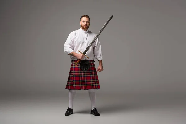 Scottish redhead man in red kilt with sword on grey background