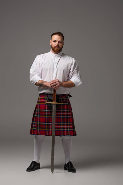 Scottish redhead man in red kilt with sword on grey background