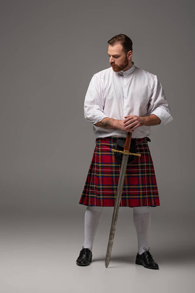 Scottish redhead man in red kilt with sword on grey background