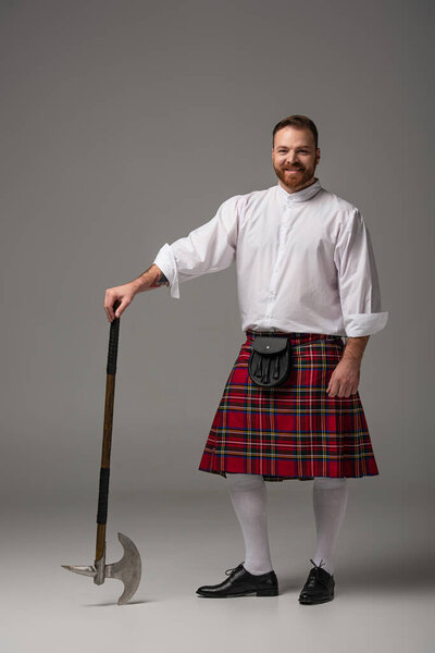 smiling Scottish redhead man in red kilt with battle axe on grey background