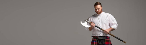 serious Scottish redhead man in red kilt with battle axe on grey background, panoramic shot
