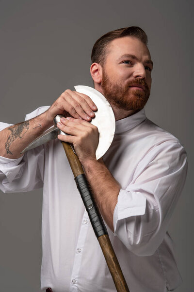 Scottish redhead man shaving beard with battle axe on grey background