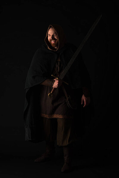 happy medieval Scottish redhead man in mantel with sword on dark isolated on black