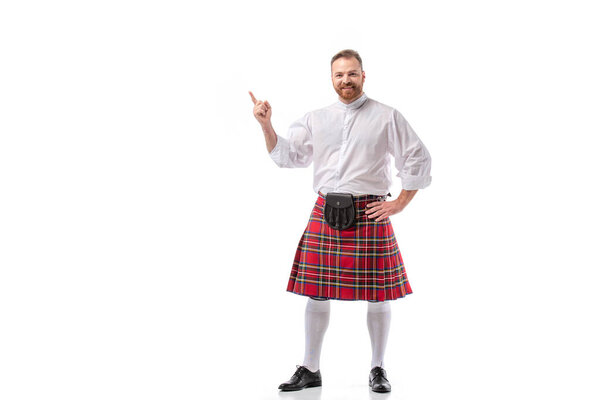 smiling Scottish redhead bearded man in red tartan kilt pointing with finger on white background