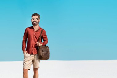 Gülümseyen resimli yüzlü adam kumlu sahilde açık mavi gökyüzüne karşı deri çanta tutuyor.