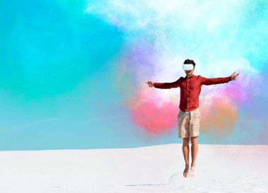 VR kulaklıklı, kumlu sahilde açık mavi gökyüzüne atlayan renkli bulutlar çizen bir adam.