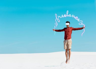 VR kulaklıklı kumsalda açık mavi gökyüzüne atlayan serbest resimli bir adam. 