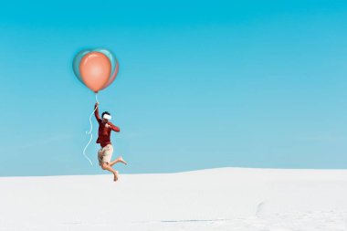 VR kulaklıklı, kumlu sahilde balonla açık mavi gökyüzüne uçan bir adam.