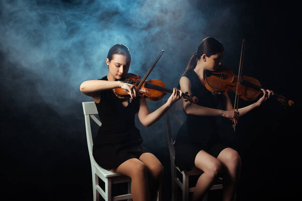 attractive female musicians playing on violins on dark stage with smoke