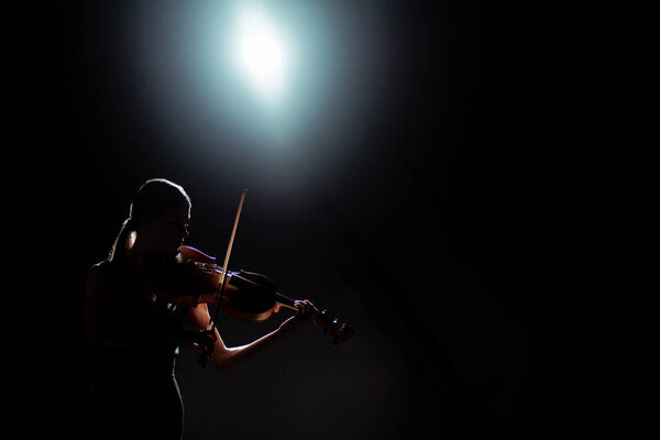 silhouette of female musician playing on violin on dark stage with back light