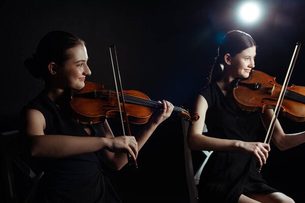 happy attractive musicians playing on violins on dark stage with back light