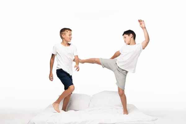 Мальчик в шутку пинает брата ногой, веселясь на кровати, изолированной на белом — стоковое фото