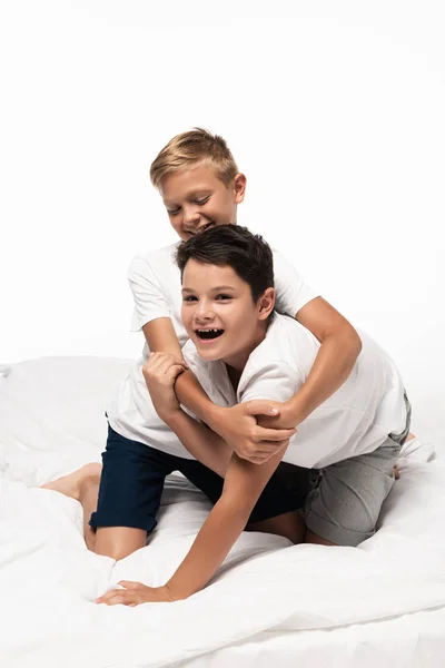 Joyeux garçon étreignant sourire frère tout en s'amusant sur le lit isolé sur blanc — Photo de stock