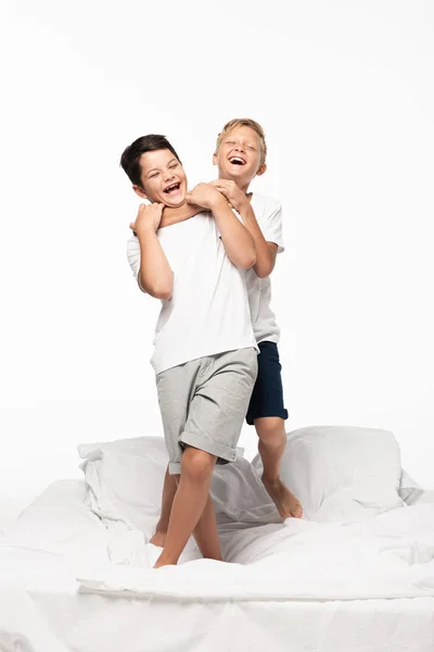 Cheerful boy jokingly stifling smiling brother while standing on bed isolated on white — Stock Photo