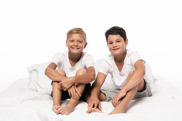 Dos hermanos alegres sentados en la cama y mirando a la cámara aislada en blanco - foto de stock