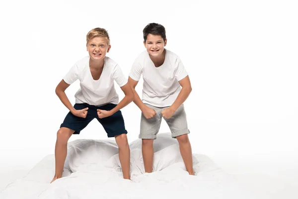 Dois irmãos alegres imitando fisiculturistas enquanto estavam de pé na cama e sorrindo isolados no branco — Fotografia de Stock