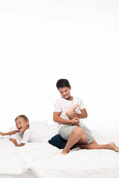 Cheerful boy jokingly breaking leg of screaming brother while having fun on bed isolated on white — Stock Photo