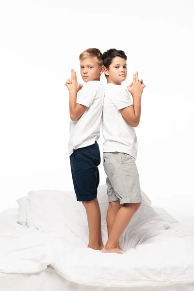 Two boys standing back to back on bed and showing gun gestures isolated on white — Stock Photo