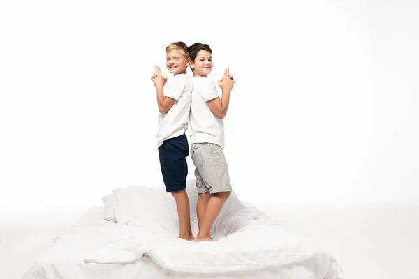 Two cheerful brothers standing back to back on bed and showing gun gestures isolated on white — Stock Photo