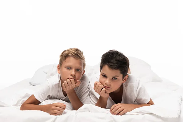 Dos hermanos pensativos acostados en la cama y sonriendo a la cámara aislados en blanco - foto de stock