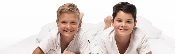 Scatto panoramico di due allegri fratelli sdraiati sul letto e sorridenti alla telecamera isolata su bianco — Foto stock