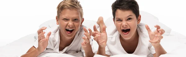 Panoramic shot of two boys grimacing and showing scaring gestures while lying on bed isolated on white — Stock Photo