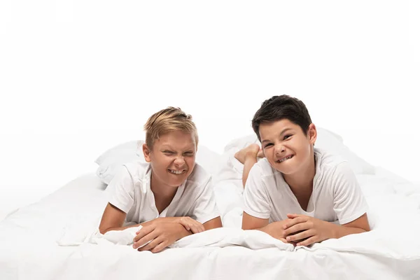 Dos chicos astutos haciendo muecas mientras están acostados en la cama y mirando a la cámara aislada en blanco - foto de stock