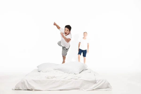 Excited boy levitating over bed while smiling brother standing on white background — Stock Photo