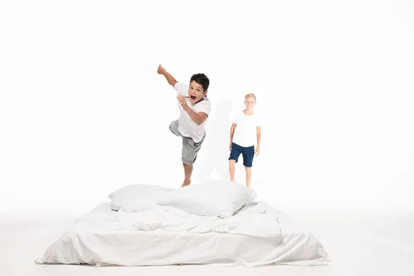 Eccitato ragazzo levitando sul letto mentre sorridente fratello in piedi su sfondo bianco — Foto stock