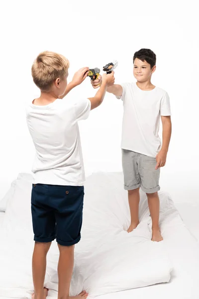 Dois irmãos apontando um para o outro com armas de brinquedo enquanto estavam de pé na cama isolados no branco — Fotografia de Stock