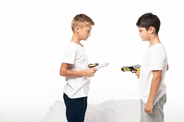 Dois irmãos apontando um para o outro com armas de brinquedo enquanto estavam de pé na cama isolados no branco — Fotografia de Stock