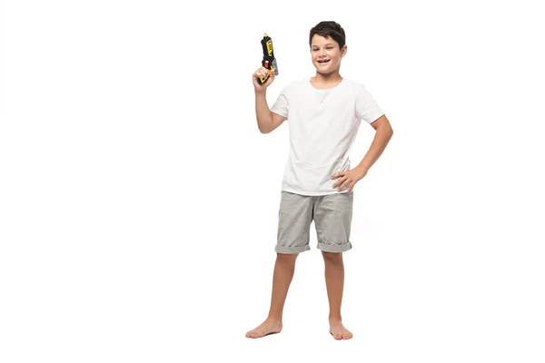 Niño alegre sosteniendo pistola de juguete mientras está de pie con las manos en las caderas y sonriendo a la cámara sobre fondo blanco - foto de stock