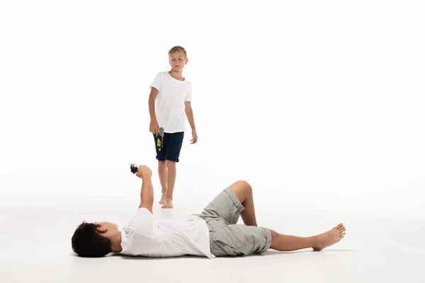 Boy pretending injured while lying and aiming with toy gun at brother on white background — Stock Photo