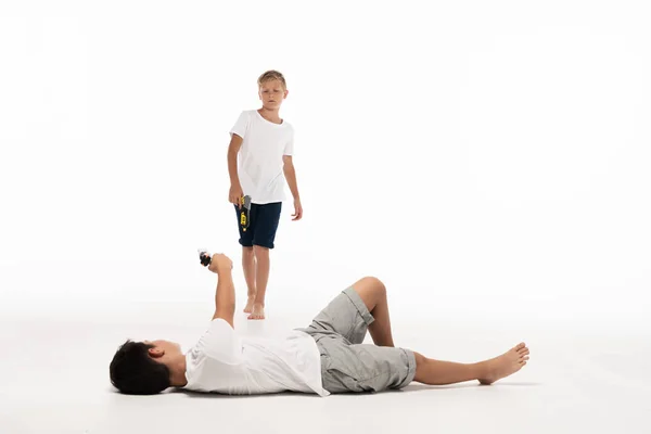 Junge täuscht Verletzung beim Liegen vor und zielt mit Spielzeugpistole auf Bruder auf weißem Hintergrund — Stockfoto