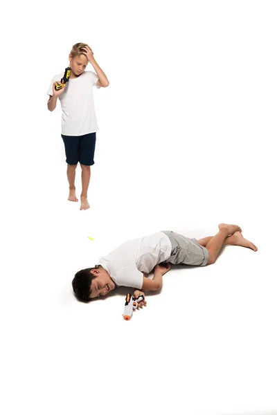 Chico fingiendo muerto cerca molesto hermano sosteniendo pistola de juguete sobre fondo blanco - foto de stock