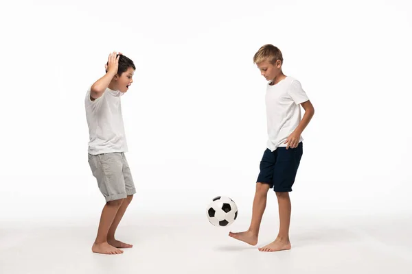 Emocionado chico mostrando wow gesto mientras mira hermano jugando con pelota de fútbol sobre fondo blanco - foto de stock