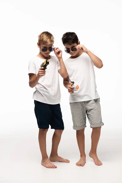 Dos hermanos en gafas de sol sosteniendo armas de juguete y mirando a la cámara mientras juegan gángsters aislados en blanco - foto de stock