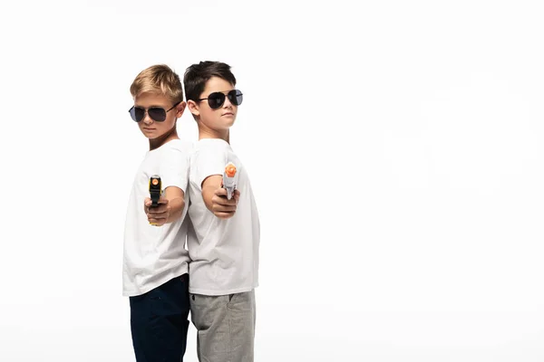 Two brothers in sunglasses standing back to back and aiming with toy guns at camera while playing gangsters isolated on white — Stock Photo