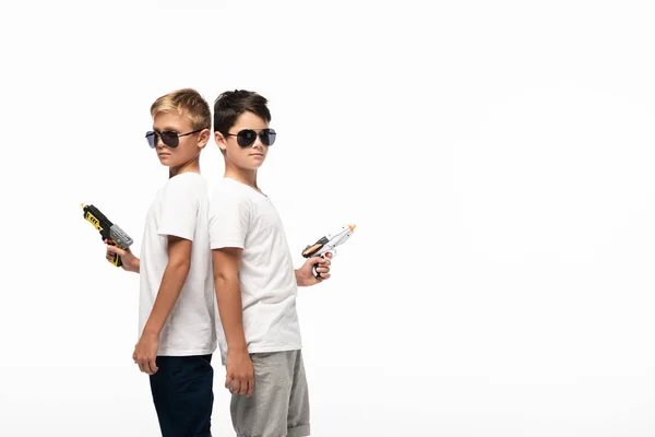 Dos hermanos en gafas de sol de pie espalda con espalda y sosteniendo armas de juguete mientras juegan gangsters aislados en blanco - foto de stock